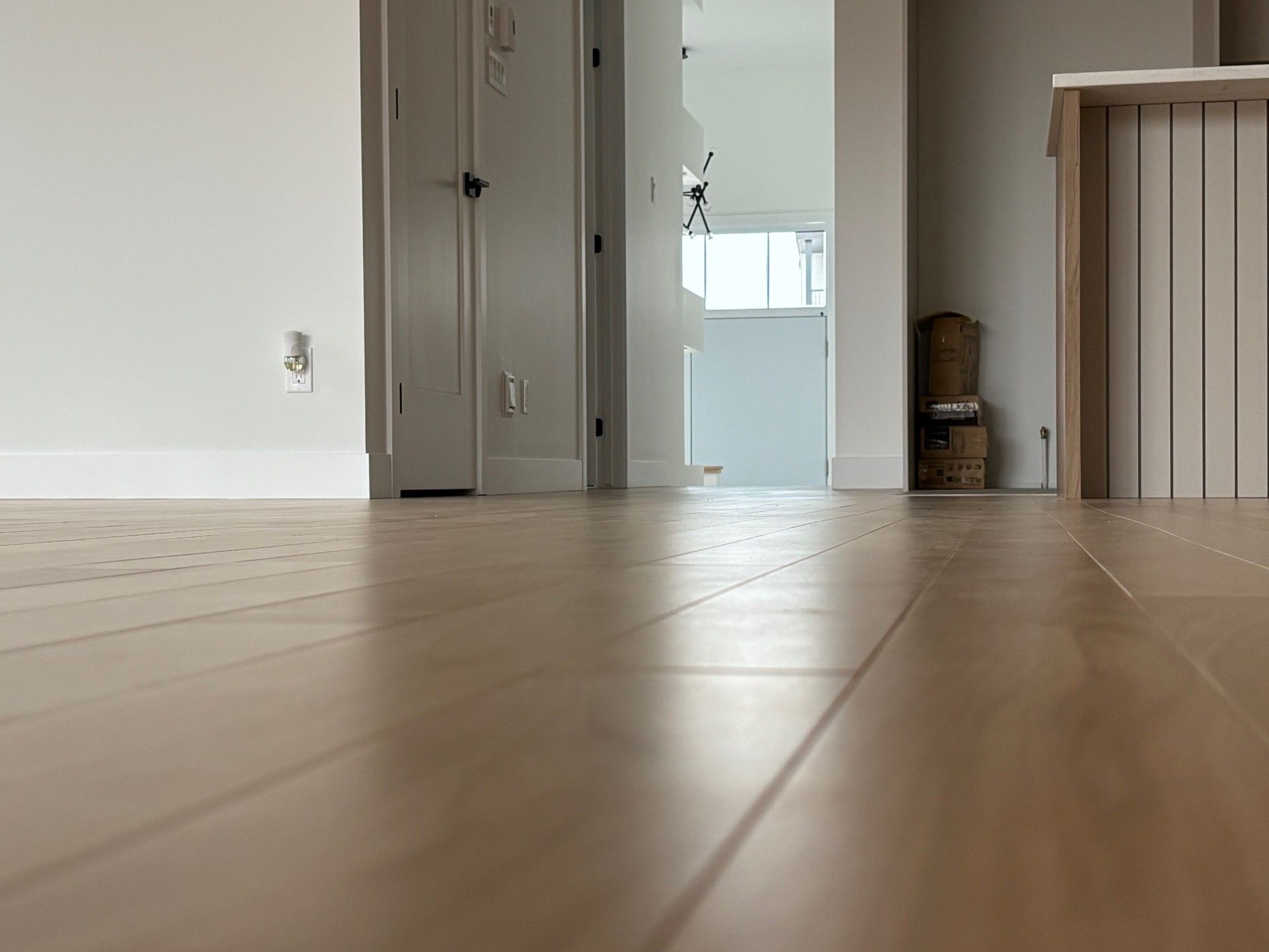 Un sol en bois mène à une porte blanche et à un couloir, avec des murs blancs et une armoire visible.