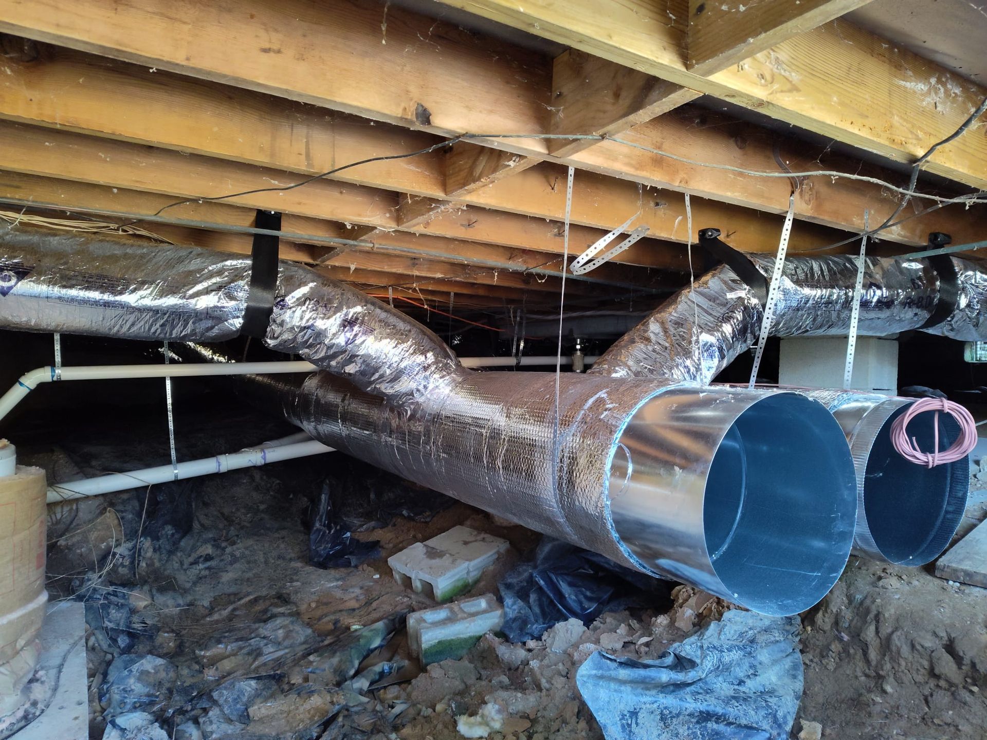 Attic with exposed ductwork and insulation beneath wooden beams and a dirt floor