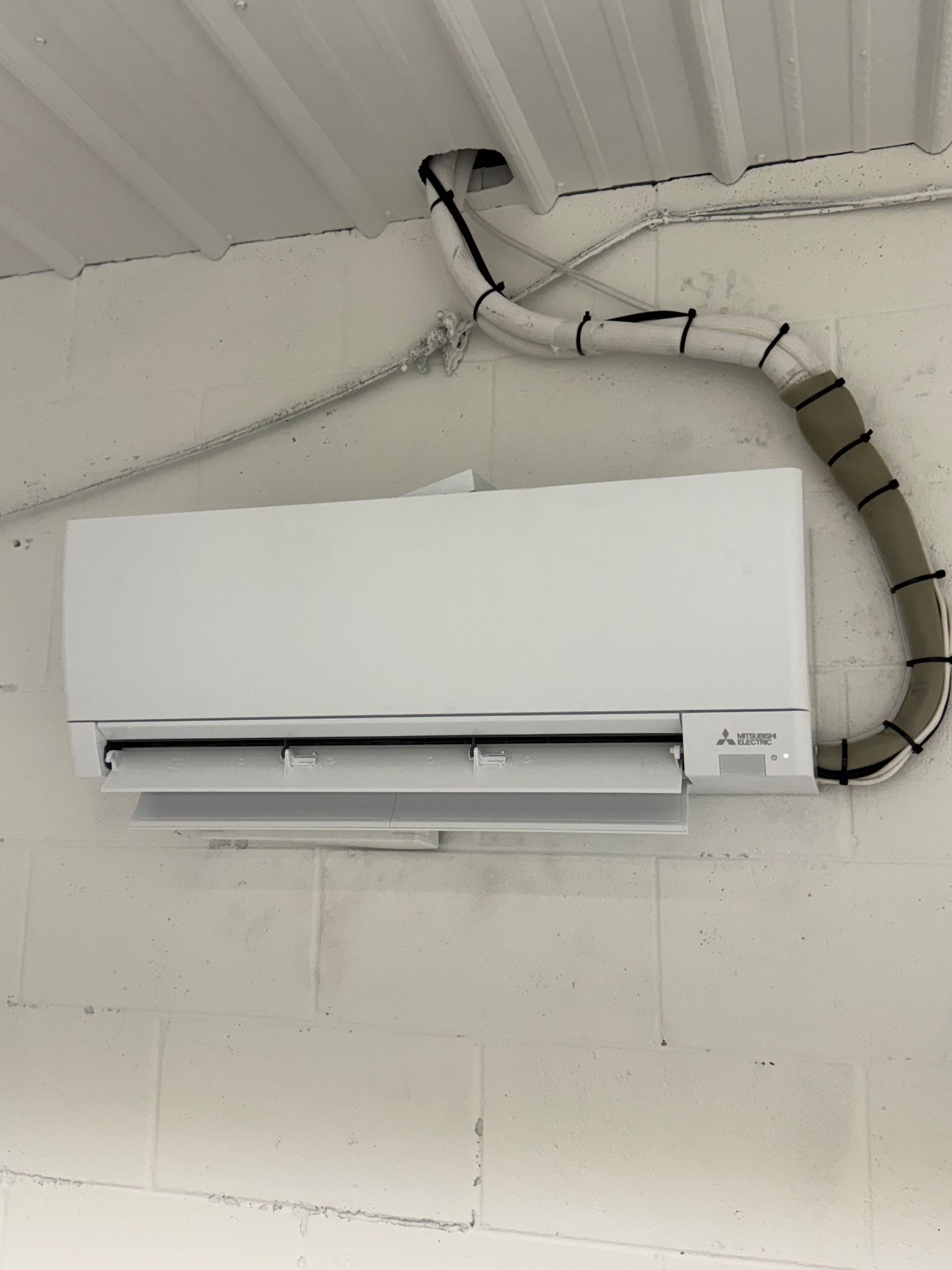 Wall-mounted white air conditioner with visible insulated tubing on a cinderblock wall.