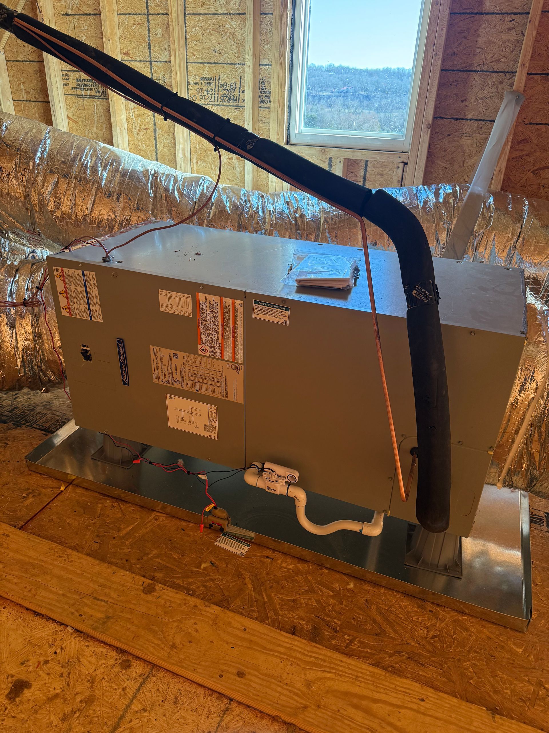 Gray HVAC unit in a room with foil-covered ducts, sitting on a wooden floor near a window.