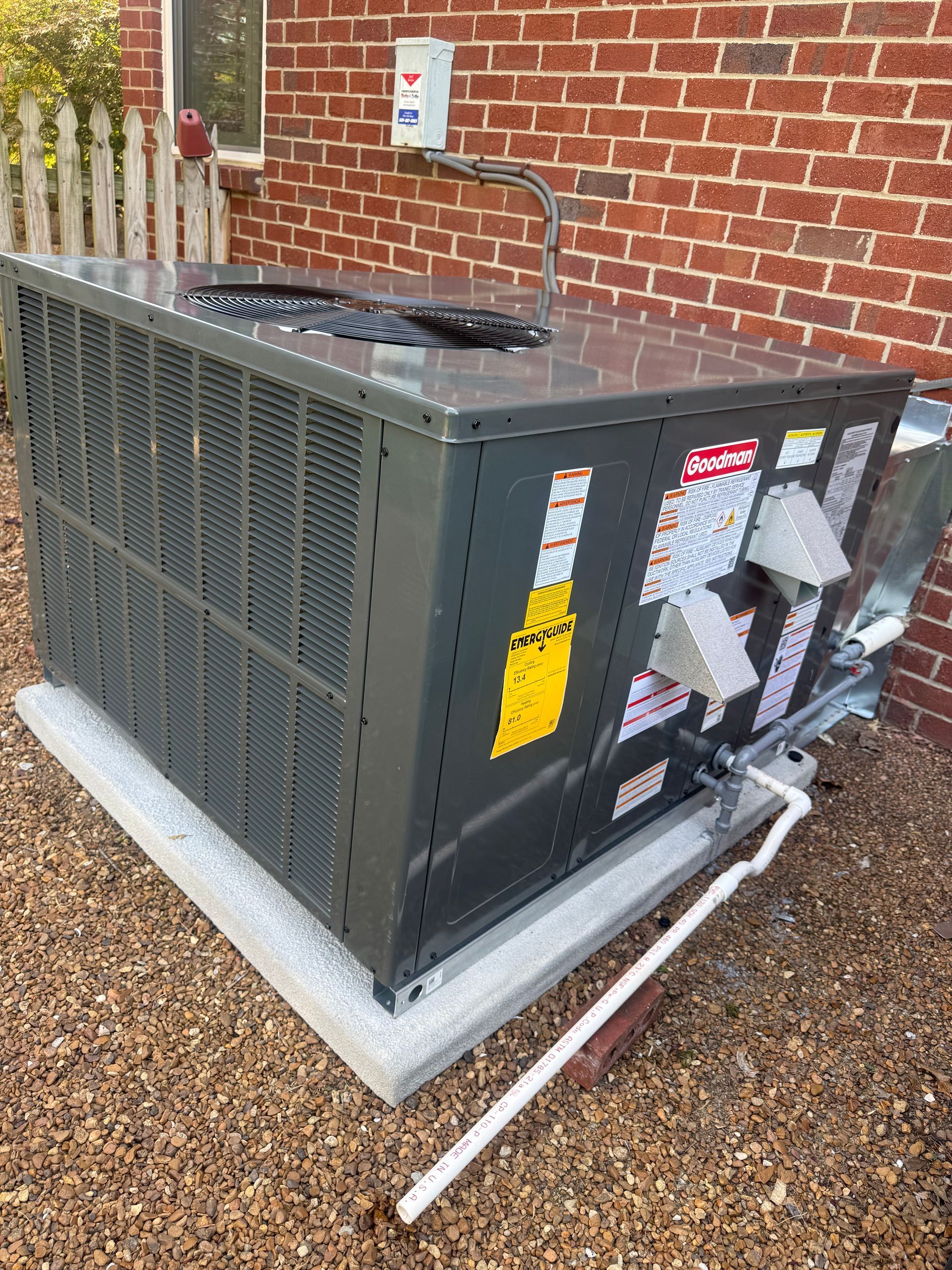 Outdoor HVAC unit on a concrete pad beside a brick wall, with exposed pipes and a white conduit on gravel.