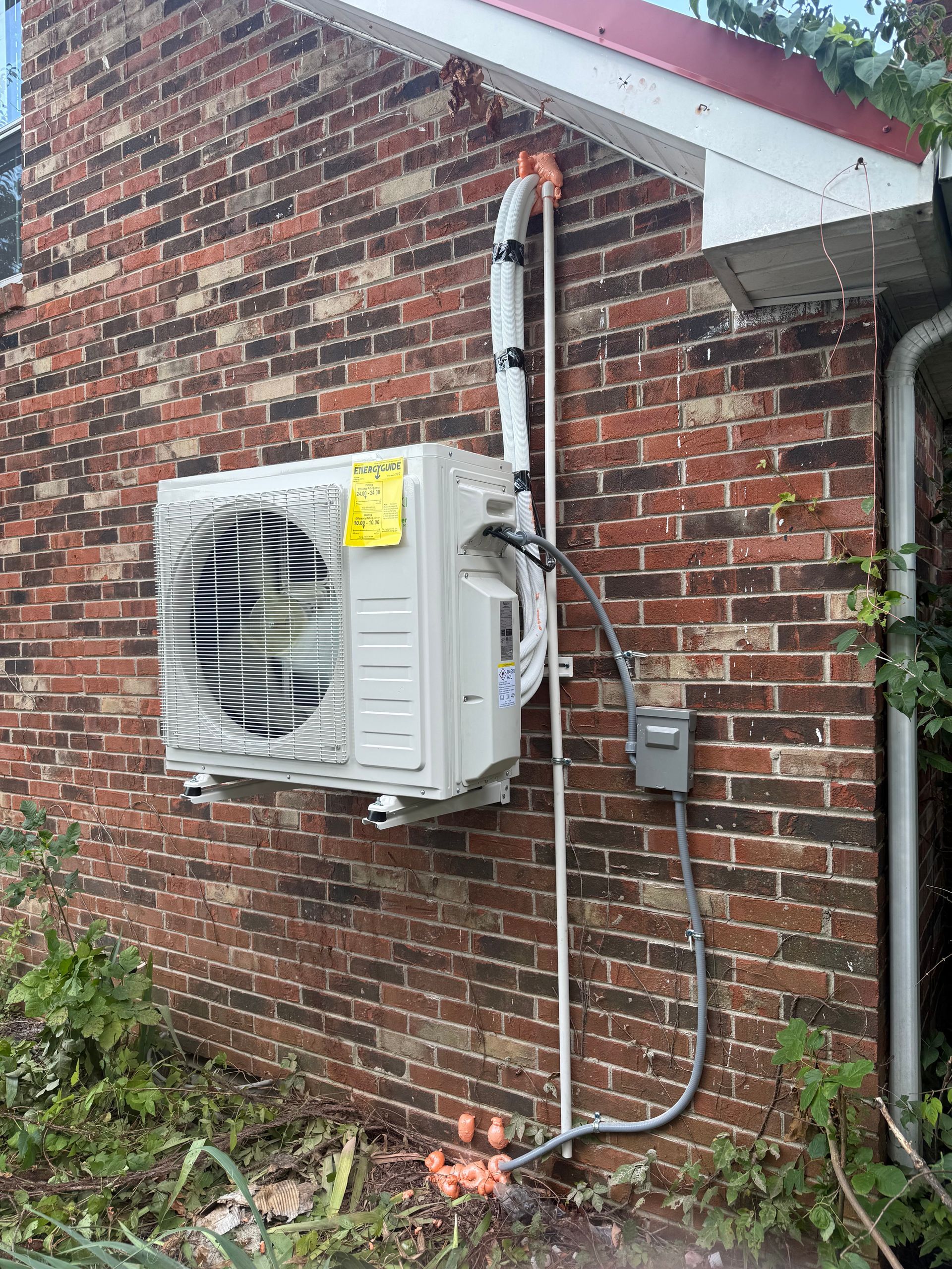 Exterior wall-mounted HVAC unit and electrical conduit on a brick house with white trim.