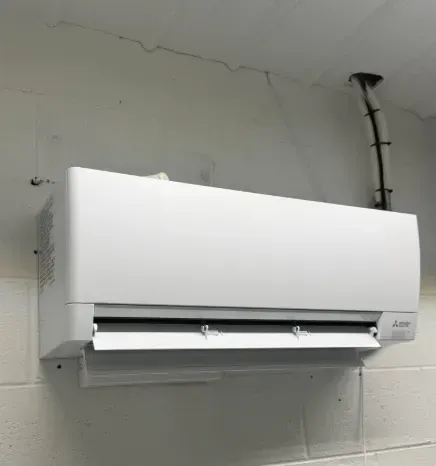 White wall-mounted HVAC unit in a bare room with exposed conduit and ceiling crack