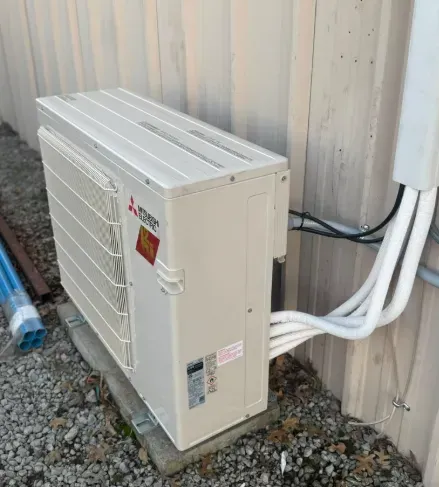 Outdoor HVAC condenser unit beside a metal wall with white pipes and gravel ground