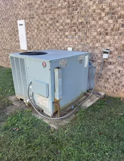Outdoor HVAC unit beside a brick wall on a concrete pad, with grass and utility lines nearby