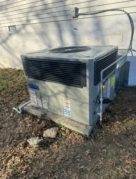 Outdoor HVAC unit beside a house, with black vents and a hose on a grassy, leaf-covered yard