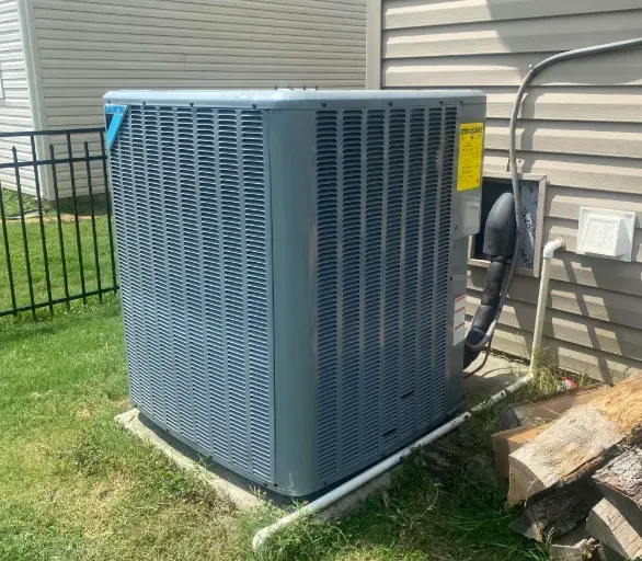 Outdoor HVAC unit beside a house with gray siding, sitting on grass near a window and patio furniture.
