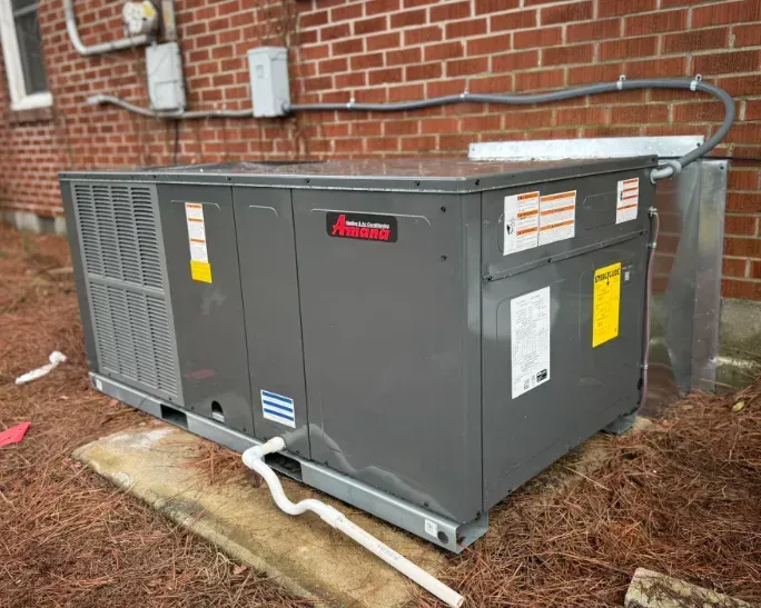 Gray outdoor HVAC unit beside a brick building, with white piping and brown mulch around it