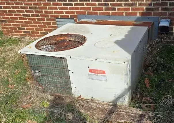 Old outdoor HVAC unit beside a brick wall, with a rusted fan grille on top.
