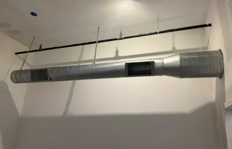 Suspended metal HVAC duct running along a white ceiling with vents and hanging supports