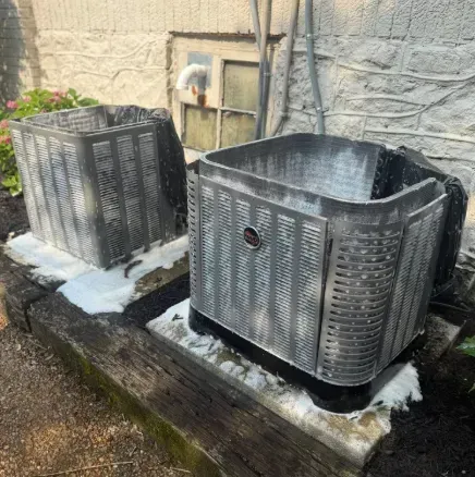 Two silver outdoor air conditioners on a concrete pad beside a stone wall, with some snow around them