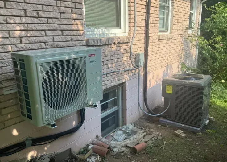 Two outdoor air-conditioning units beside a stone house, with a window and garden debris nearby
