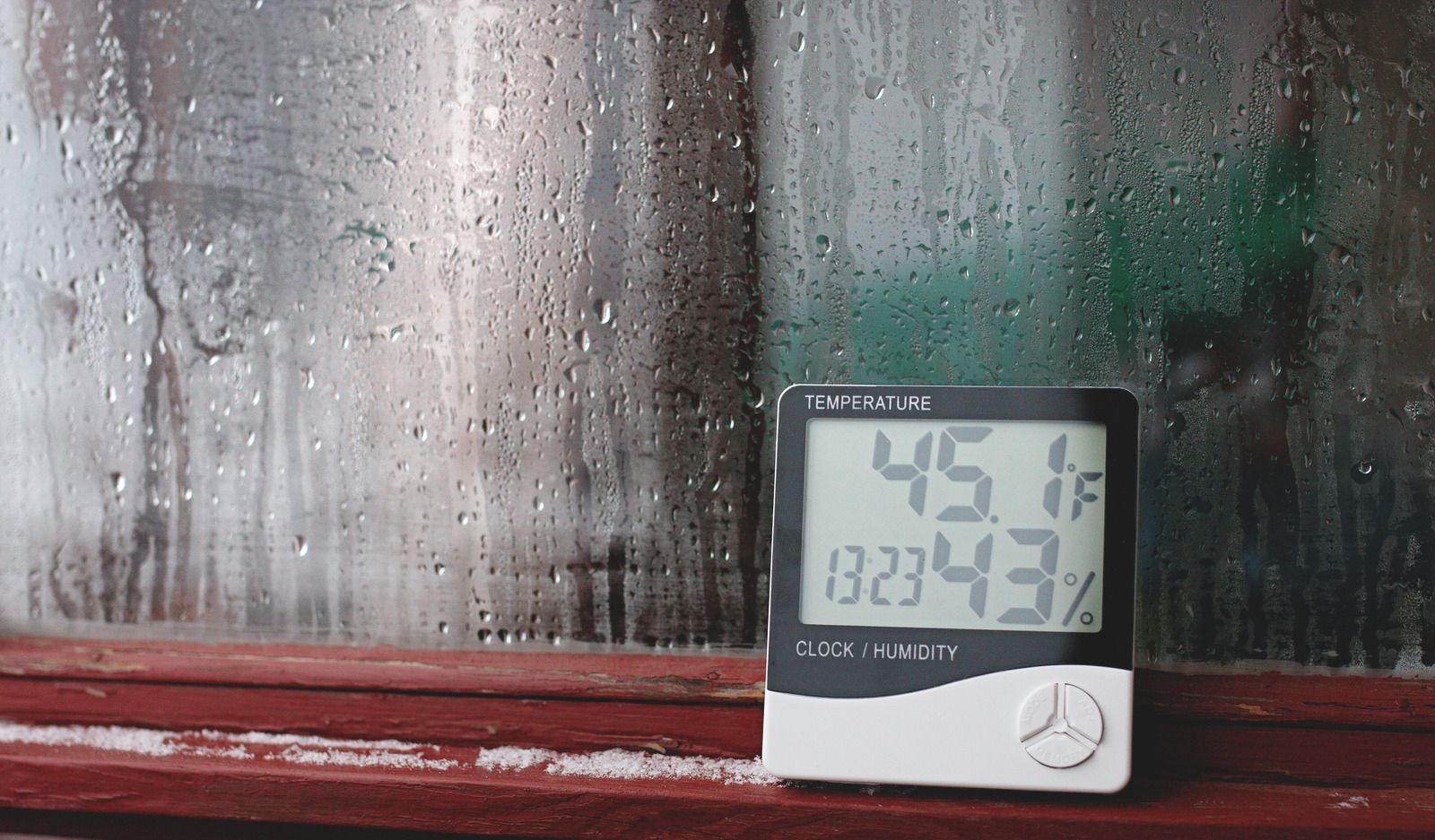 Small digital thermometer/hygrometer on a windowsill beside a wet, streaked window showing 45.5 and 12:43.