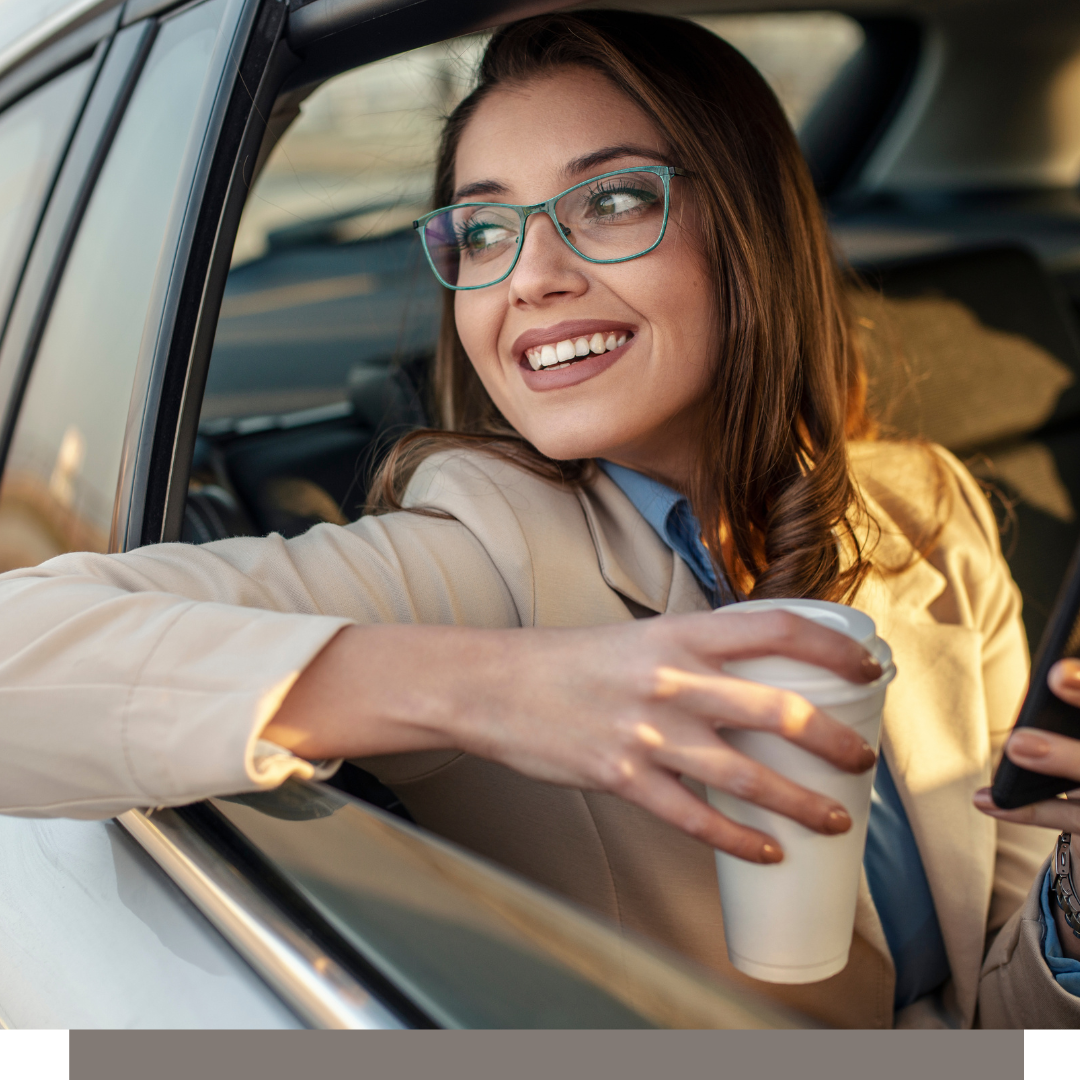 Smiling woman in glasses leans out a car window holding a takeaway coffee cup