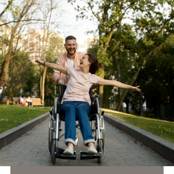 Man pushing a person in a wheelchair along a park path, both smiling with arms outstretched.