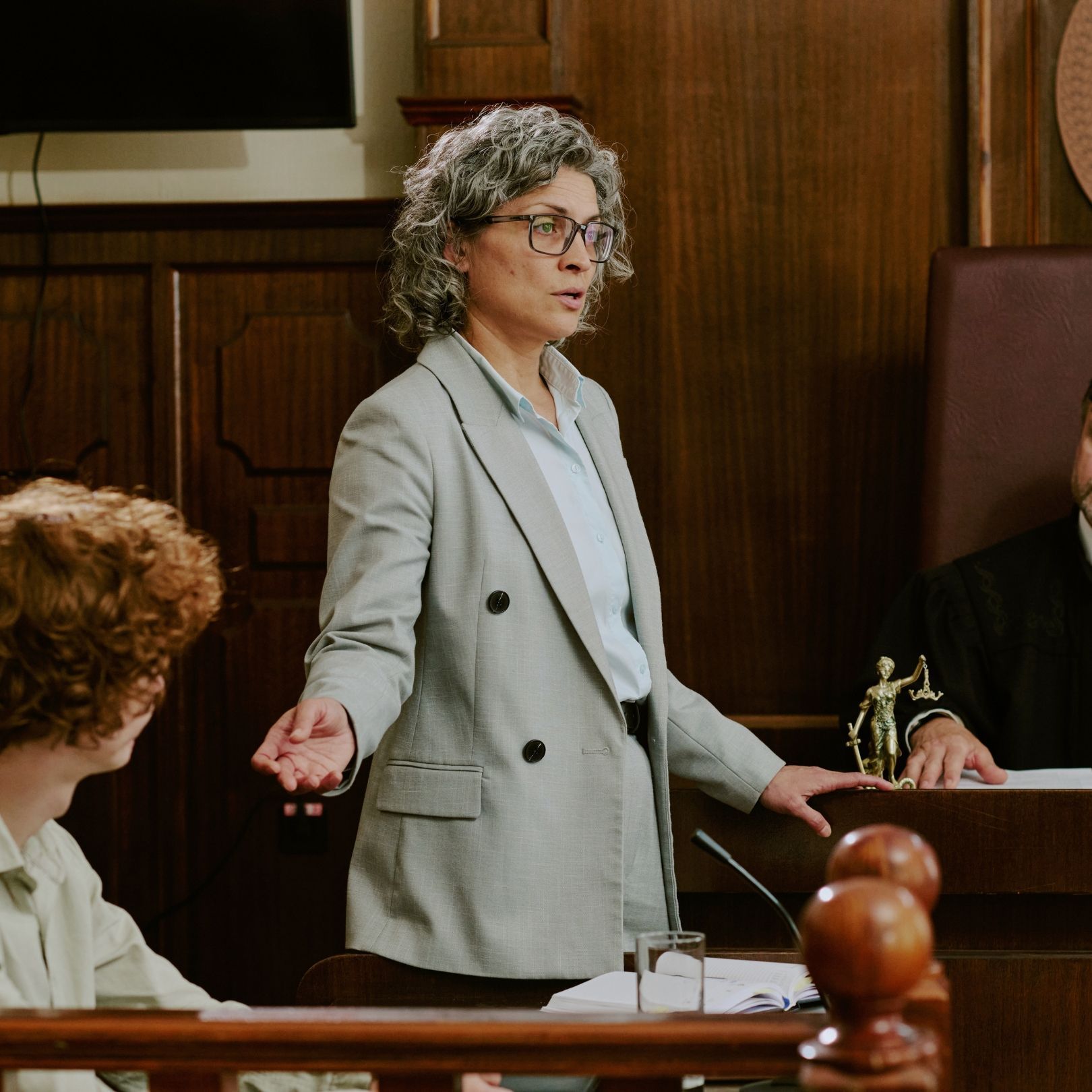 Woman in gray suit speaking in a courtroom, standing beside the bench with one hand raised.