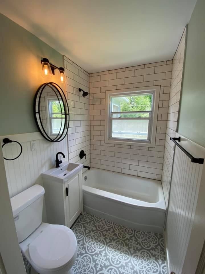 Small bathroom with white tub, toilet, pedestal sink, oval mirror, and patterned gray floor tiles
