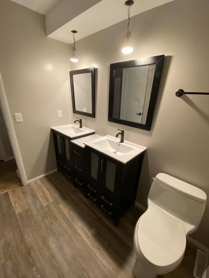 Modern bathroom with double vanity, large mirrors, and a toilet on wood-look flooring.