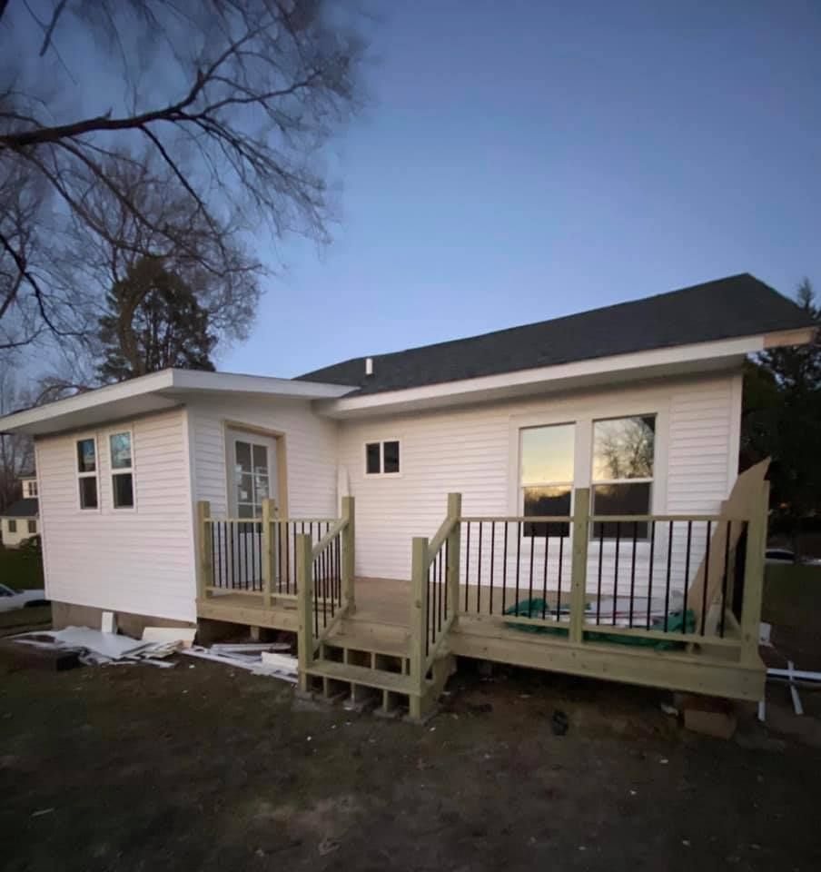 Small beige house with a deck and steps, photographed at dusk in a yard with bare trees.
