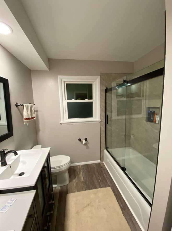 Small bathroom with white vanity, toilet, window, and glass-enclosed shower over gray wood-look floor.
