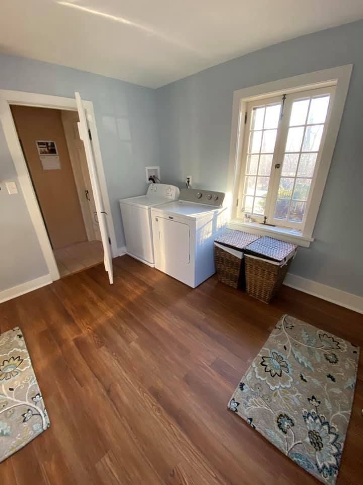 Laundry room with white washer and dryer, wood floor, window, and wicker basket beside a small rug.