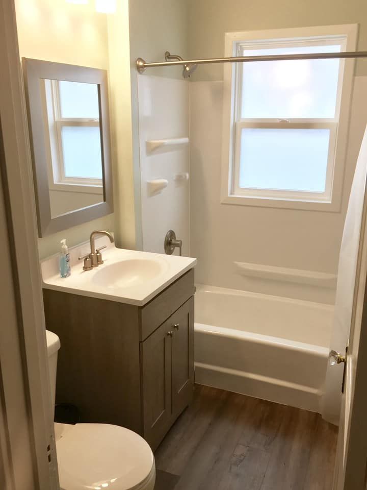 Small bathroom with white tub, vanity, toilet, mirror, and window.