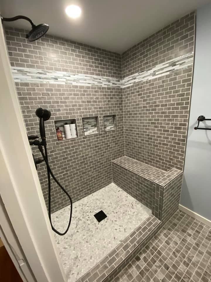 Small tiled walk-in shower with brick-pattern walls, built-in shelf, bench, and black showerhead.