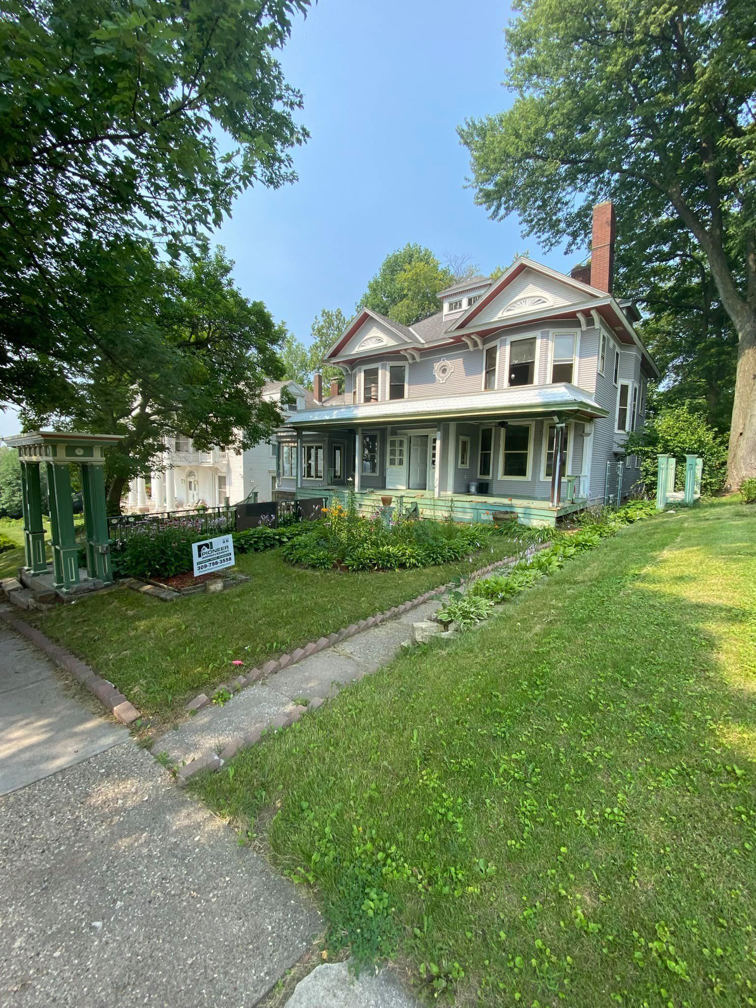 Victorian house with white porch and green lawn, shaded by trees along a sidewalk
