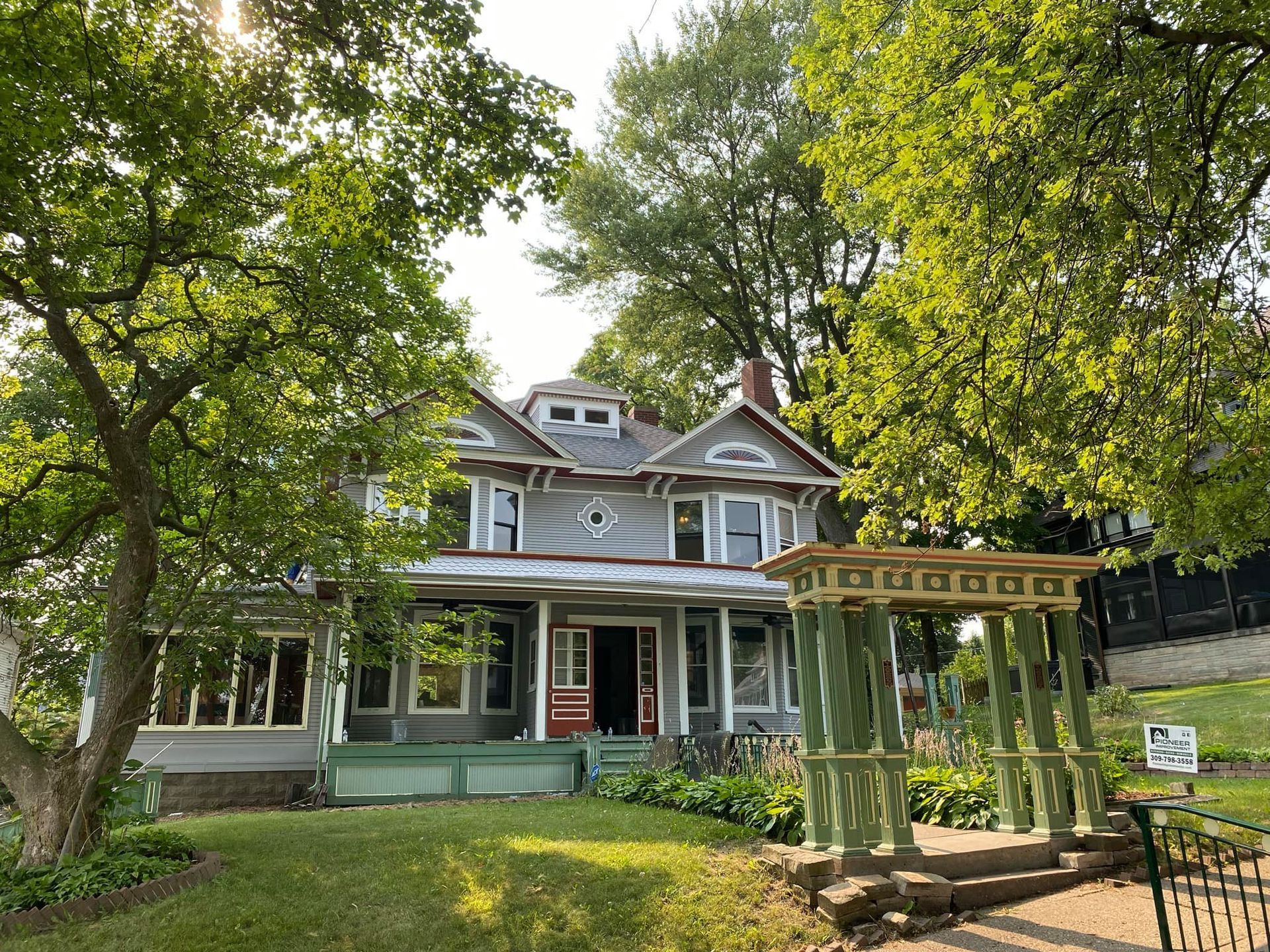 Victorian house with green porch and pergola, surrounded by trees in a sunny yard