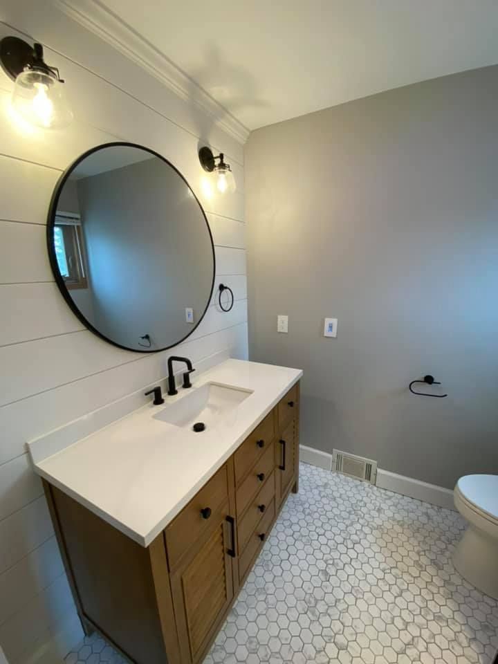 Modern bathroom with round mirror, wood vanity, white countertop, gray walls, and hex tile floor