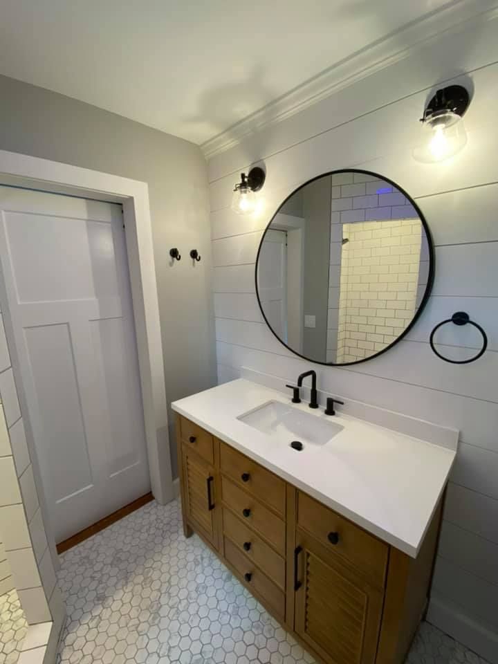 Bright bathroom vanity with round mirror, white sink, black fixtures, and wood cabinet against light walls.