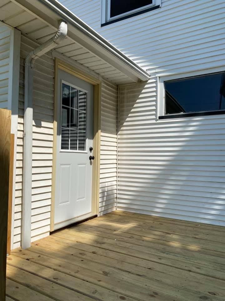 White back door under a small porch beside beige siding and a sunlit wooden deck