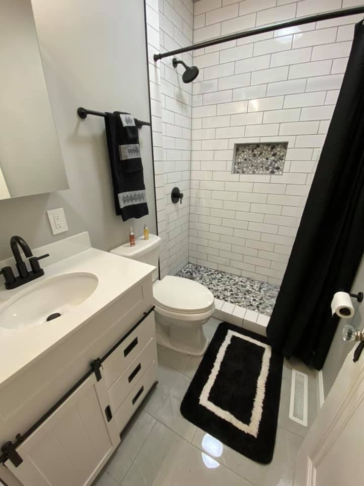 Small modern bathroom with white tile, black accents, shower, toilet, sink, and striped rugs.