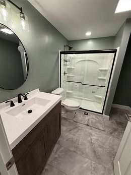 Modern bathroom with dark vanity, white sink, round mirror, gray tile, and glass-enclosed shower.