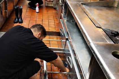Learn More Two people in black outfits clean commercial kitchen one scrubs drawer, other cleans cabinet, showing focus and teamwork.