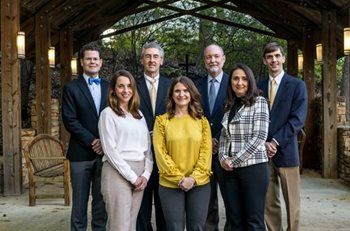 Seven people standing together in a rustic wooden barn, dressed in business-casual attire, smiling at the camera