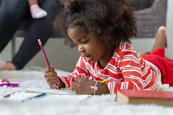 Child drawing on paper with a red pencil while lying on the floor in a red striped shirt