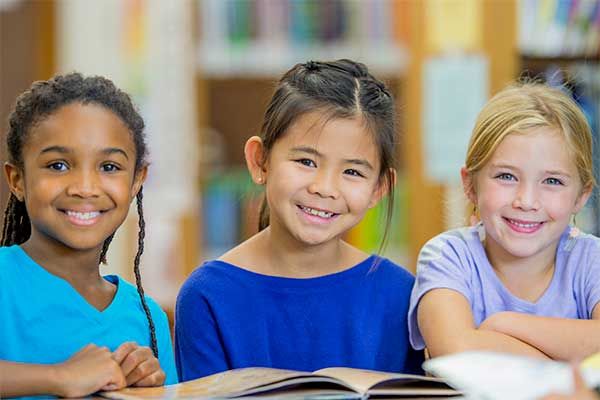 Three children smiling over an open book in a classroom setting