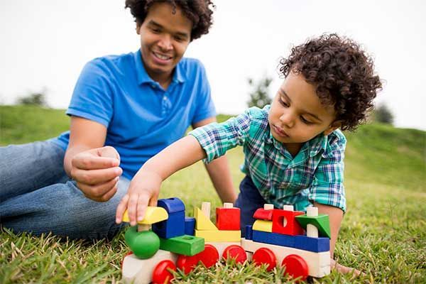 Adult and child playing with colorful blocks and toy vehicles on grass outdoors