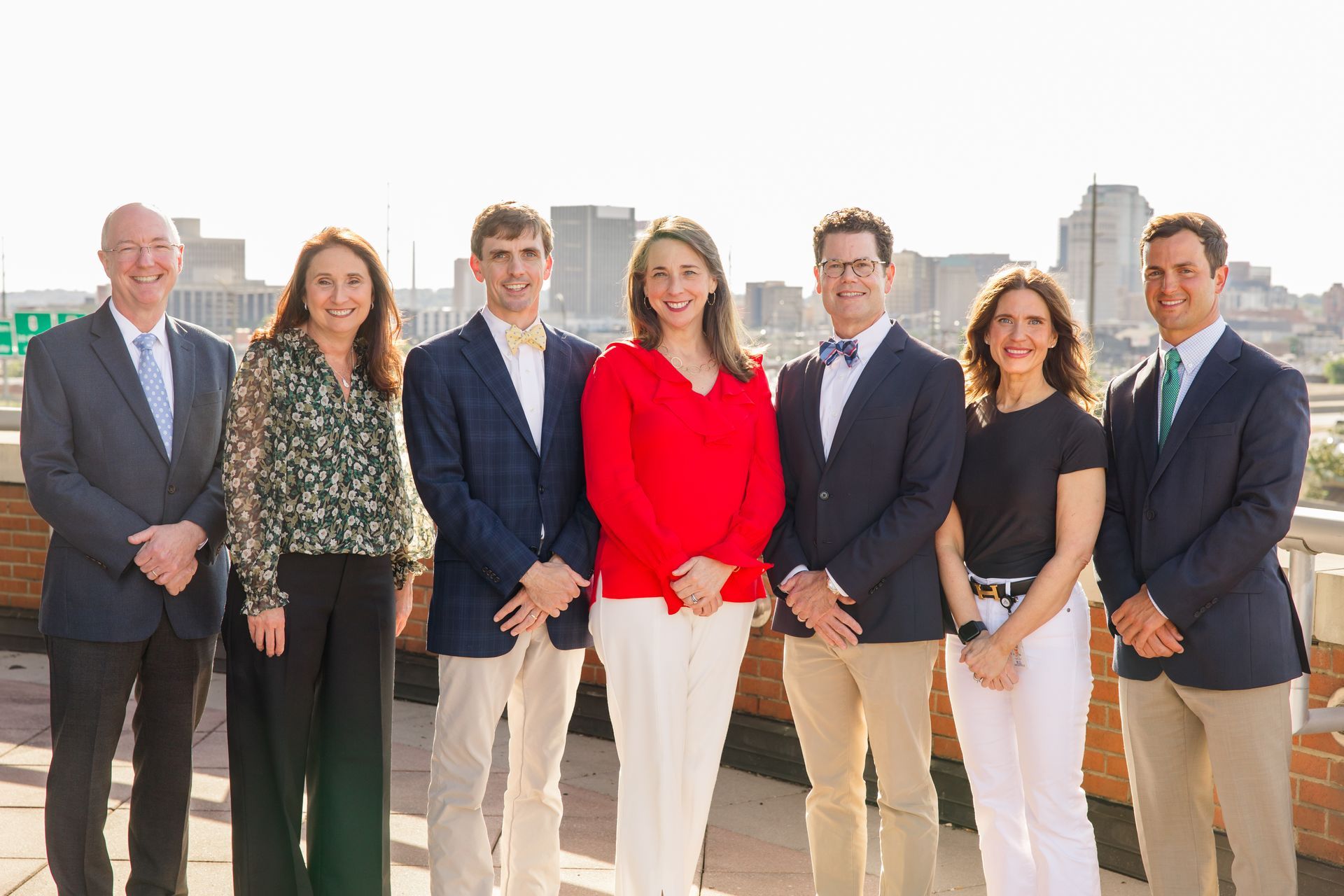 Seven people posing on a rooftop terrace with city skyline in the background, wearing business attire.