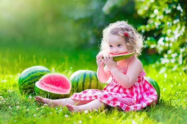 Child in a pink dress eating watermelon on grass beside whole and sliced watermelons