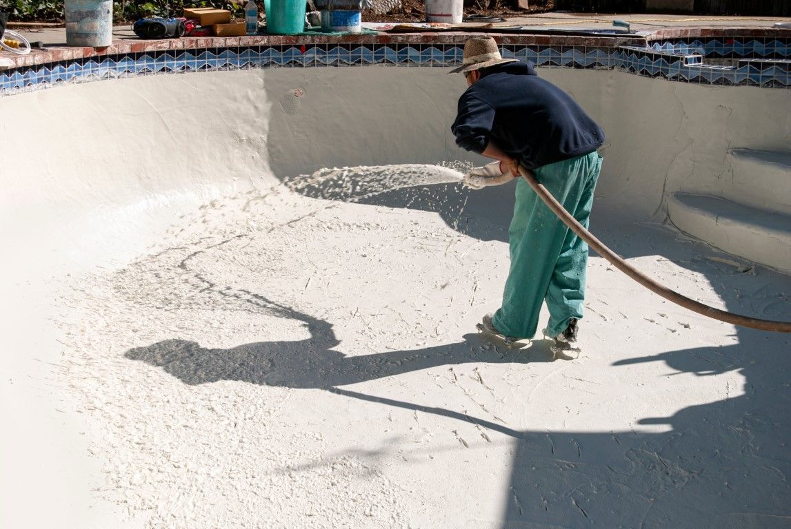 Worker spraying white coating inside an empty concrete pool with a hose