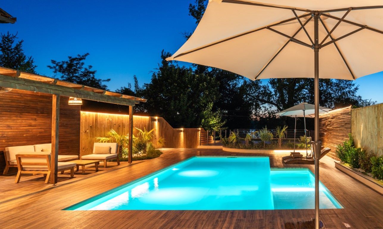 Modern backyard pool at dusk with wooden deck, lounge chairs, and a large patio umbrella