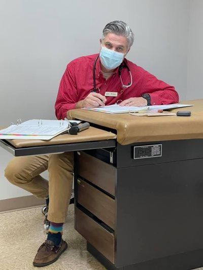 A person in a red shirt and face mask sits at a desk, writing. They wear colorful socks and have a stethoscope.