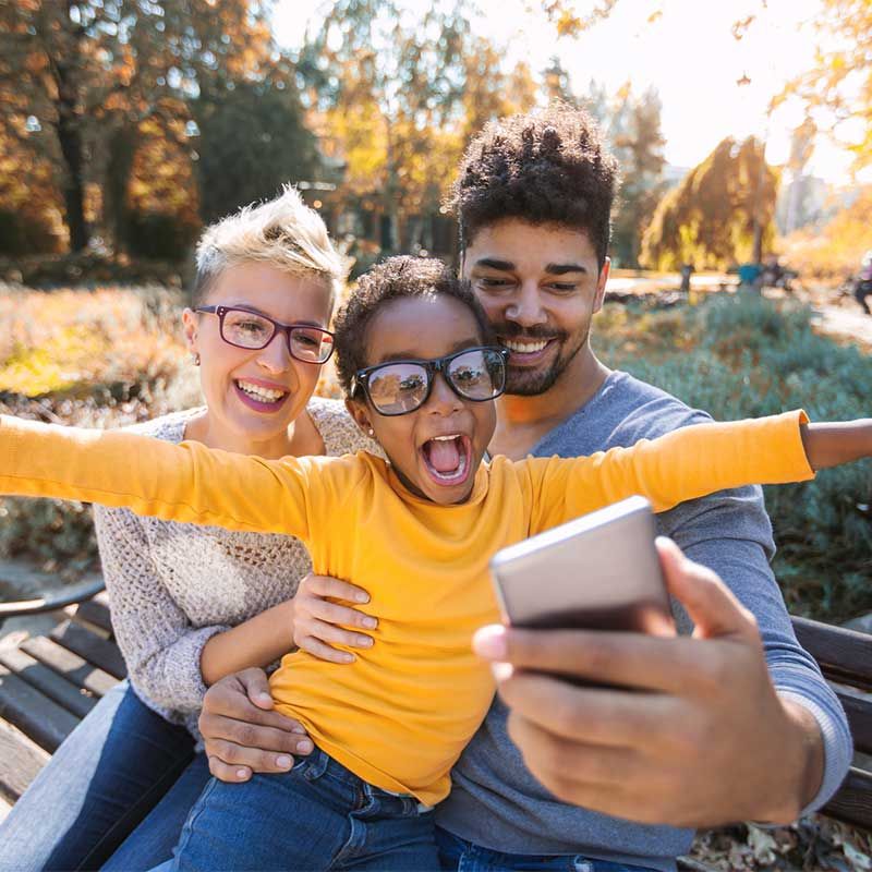Family taking a selfie outdoors; child in yellow shirt with arms outstretched, smiles with parents on a bench.