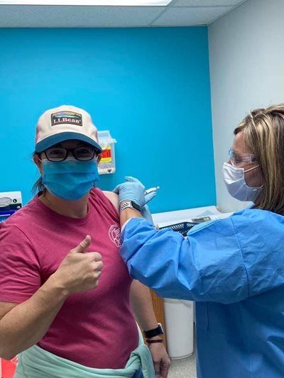 Woman in a pink shirt getting a shot in the arm; gives a thumbs up. A healthcare worker administers in a blue gown.