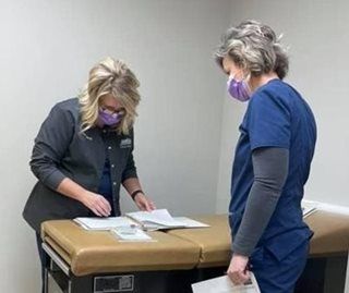 Two masked medical workers review paperwork near an examination table in a medical office.