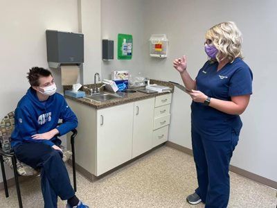 A healthcare worker in scrubs explains information to a person seated in a doctor's office. Both wear masks.