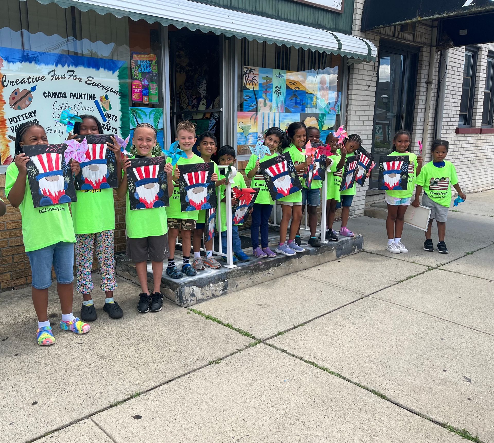 A group of children in bright yellow shirts stand in front of an art studio holding handmade patriotic star signs.