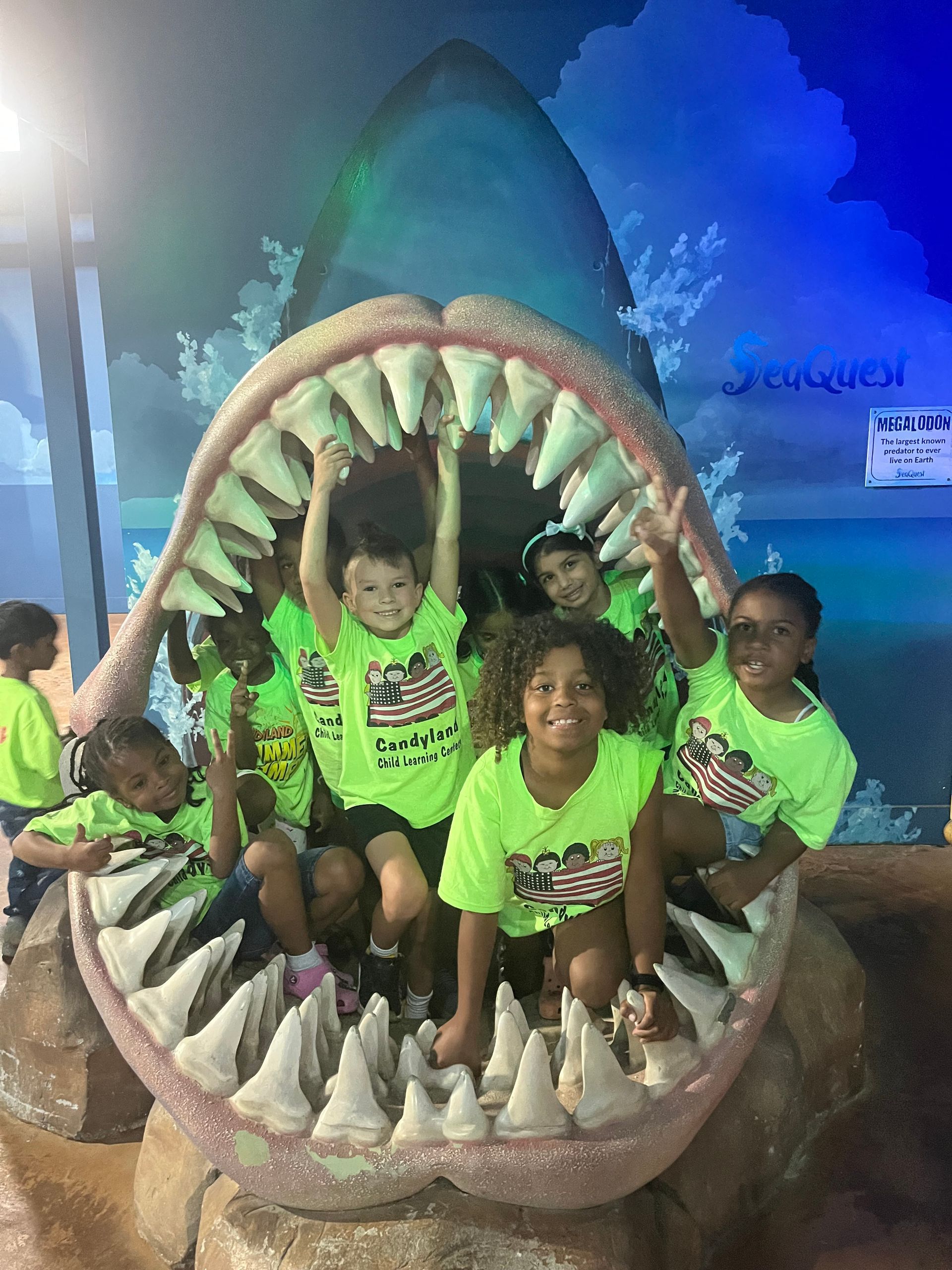 Group of children in matching neon green shirts posing inside a large shark jaw sculpture in a blue-lit indoor setting.